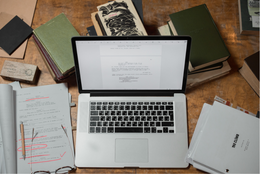 nonfiction laptop Desktop with laptop in the center surrounded by scattered books, scripts, glasses, and stationary supplies representing research for nonfiction writing