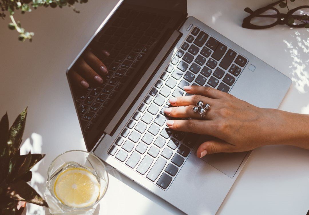 Woman with tablet and laptop A woman's hand on the keyboard of a laptop with plants, reading glasses, and a glass of lemon water in the periphery, representing a romance author preparing manuscript for submission